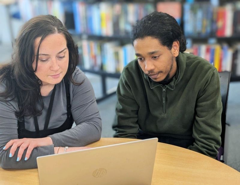 Two people seated at a table in a library or learning space, looking at an open laptop together. Bookshelves filled with books are visible in the background, and the table occupies the foreground.