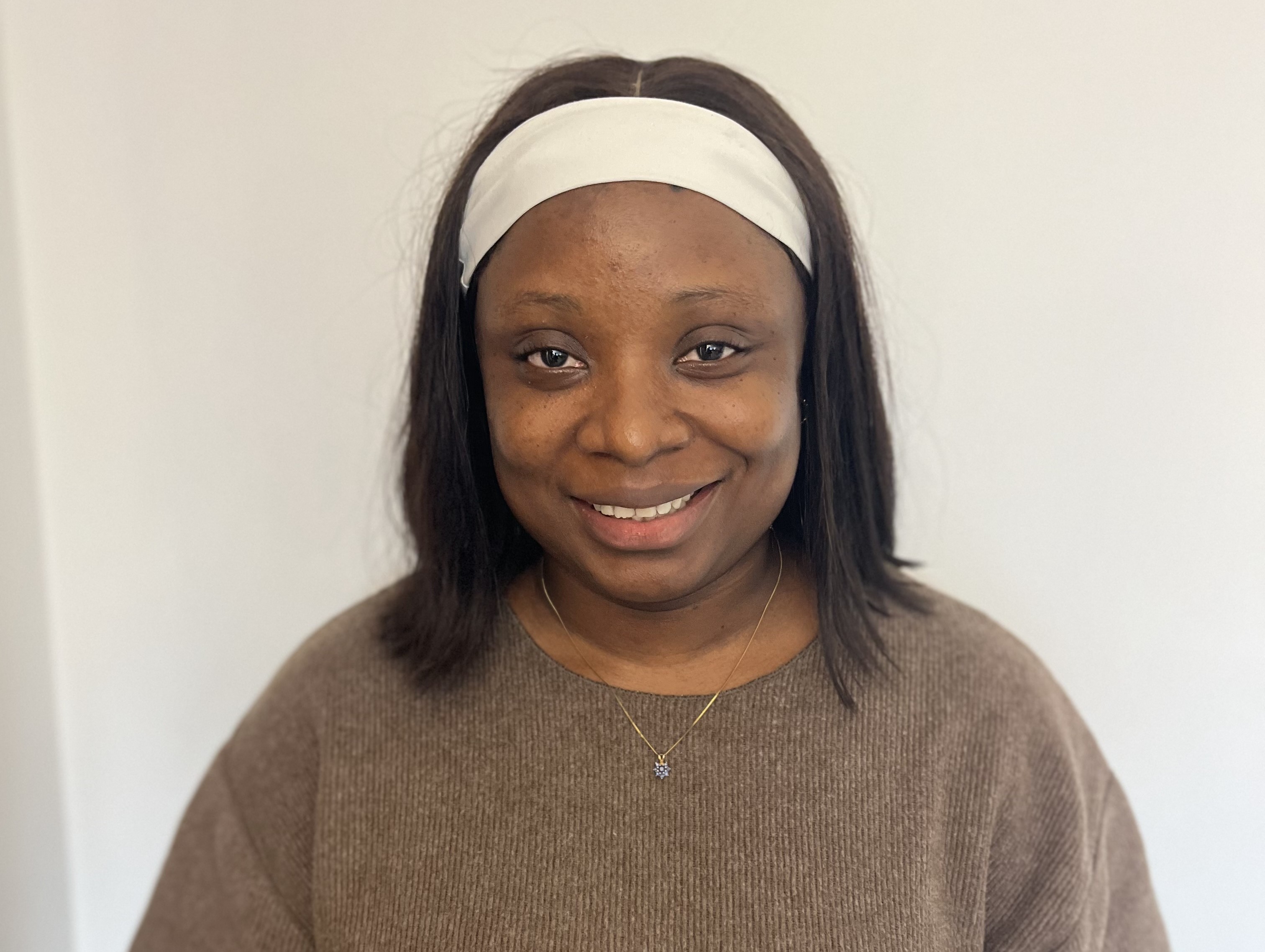 A woman stands smiling against a white wall. She is wearing a white headband and brown knitted jumper.