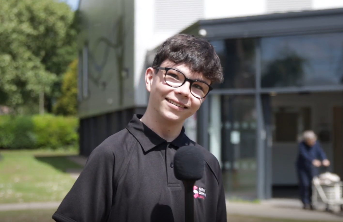 A young man wearing glasses and a black polo shirt stands smiling in front of a building and trees