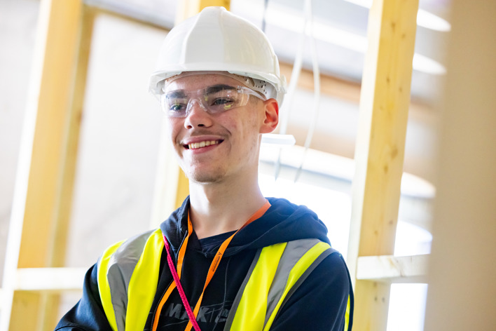 A young man wearing a white hard hat, safety glasses, a navy blue hoodie, orange lanyard and bbight green hi-vis jacket stands smiling. In the background, wooden joists can be seen.