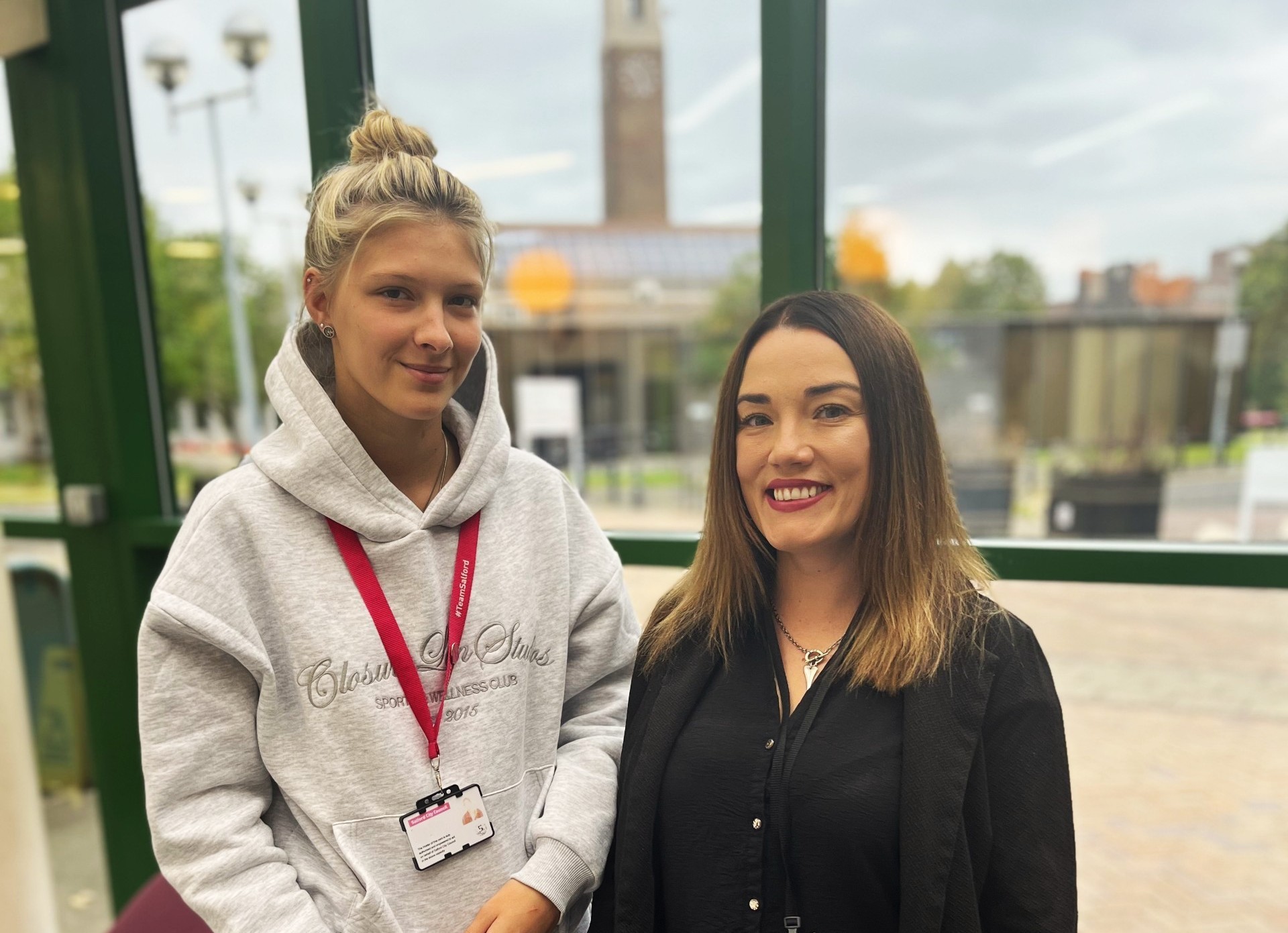 Two women stand smiling in front of a window. In the background, a clock tower can be seen out of focus.