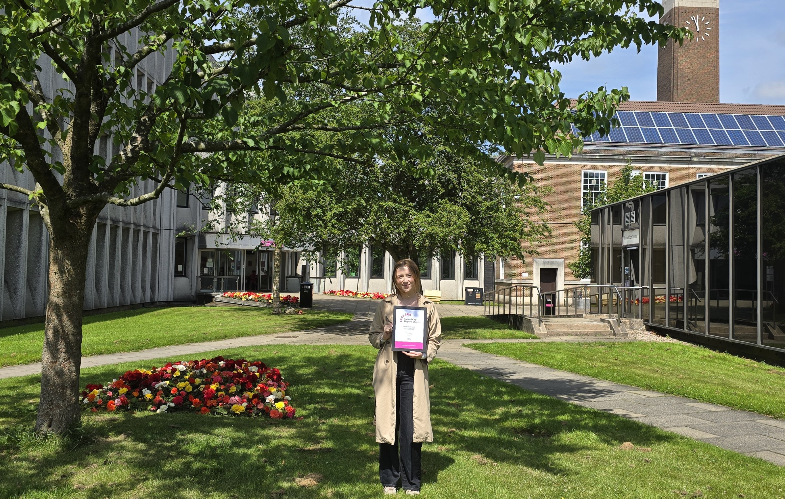 A woman stands outdoors smiling and holding a framed certificate. She is wearing a beige coloured long coat and black trousers. In the background there are trees, flowers, and various buildings including a clock tower.
