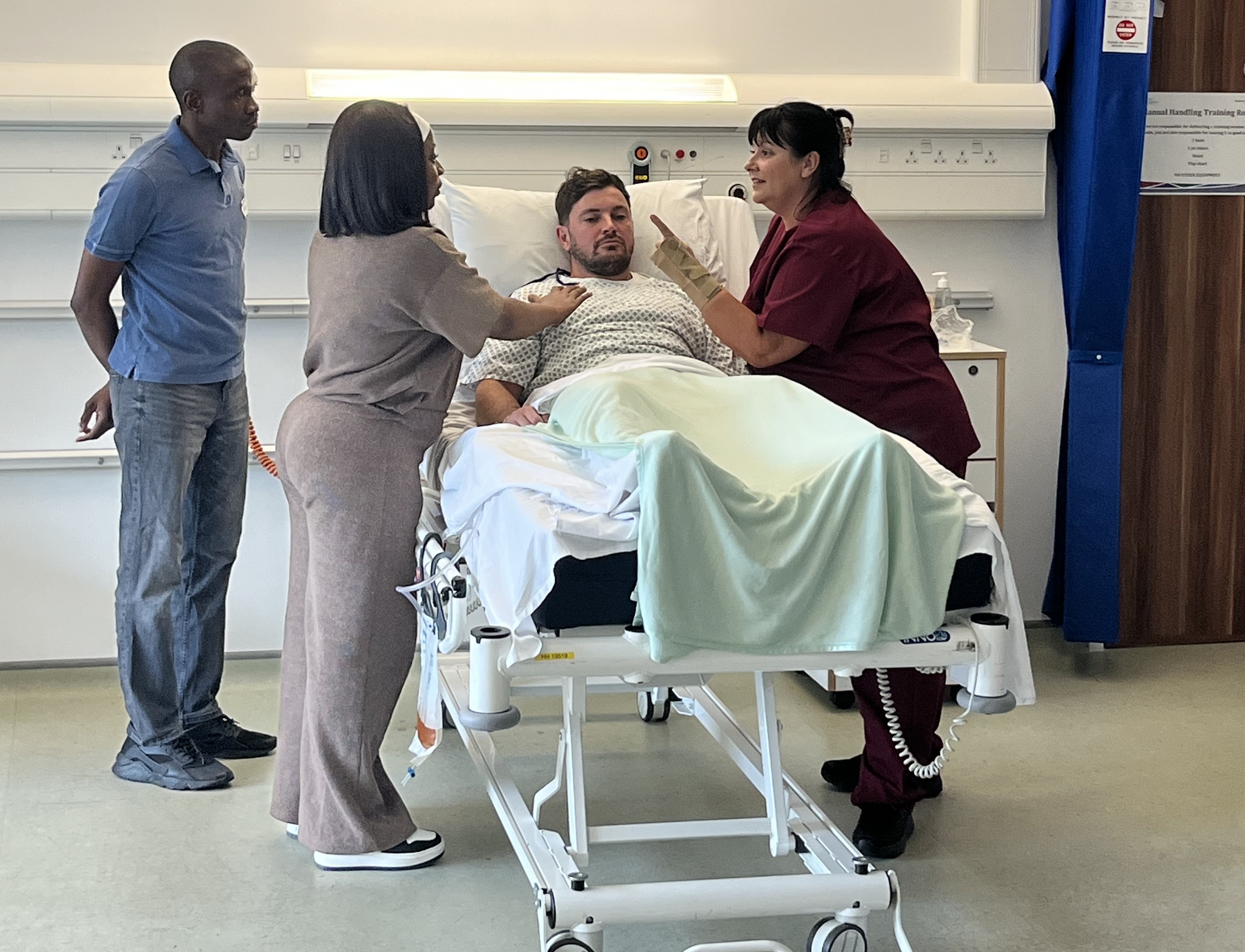 A man lies in a hospital bed with a man and two women stood surrounding the bed having a conversation.