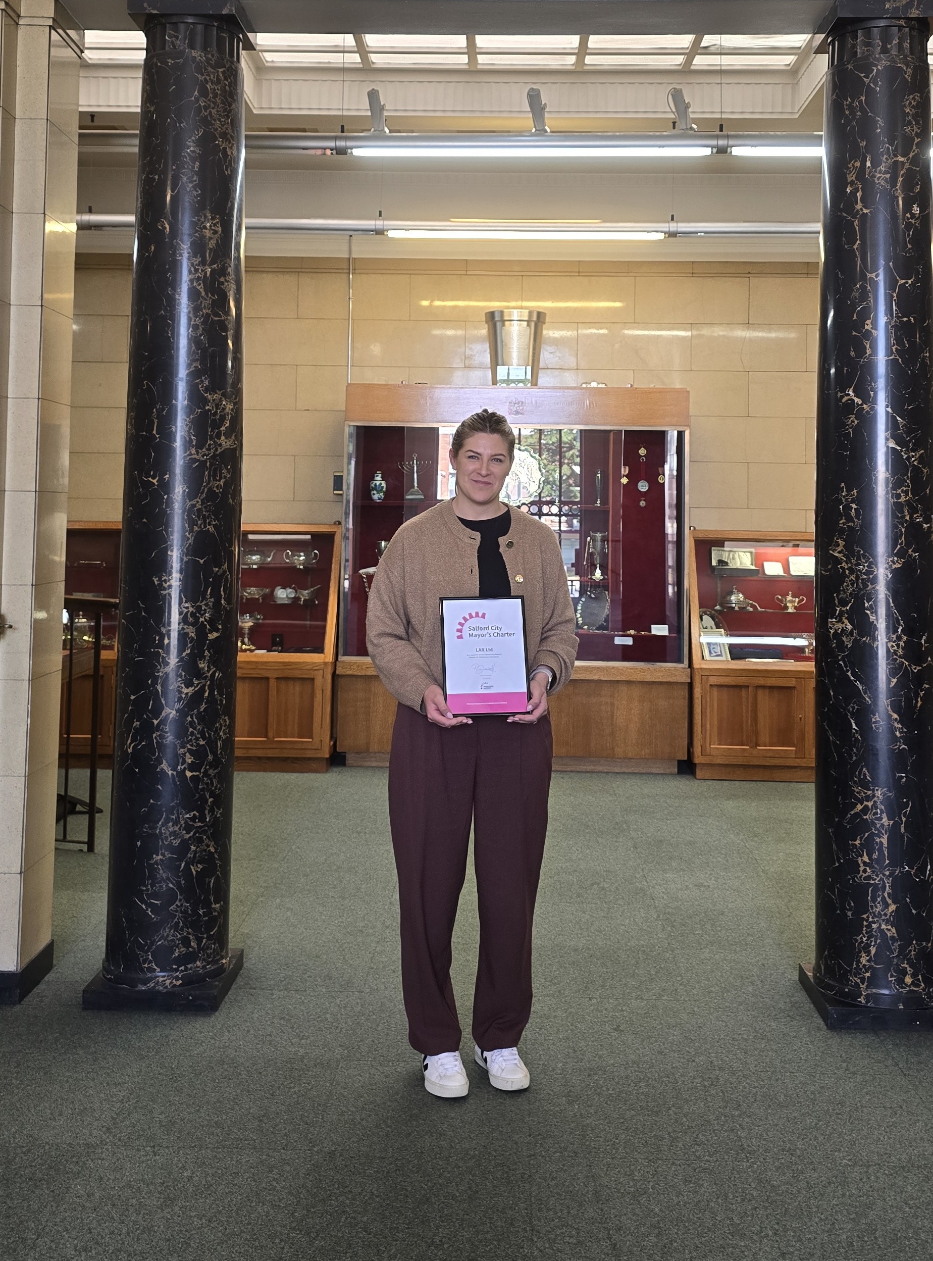 A woman standing smiling and holding a framed certificate. She is wearing a brown cardigan and black trousers with white trainers. In the background, two black pillars are visible, as well as some wooden and glass cabinets.