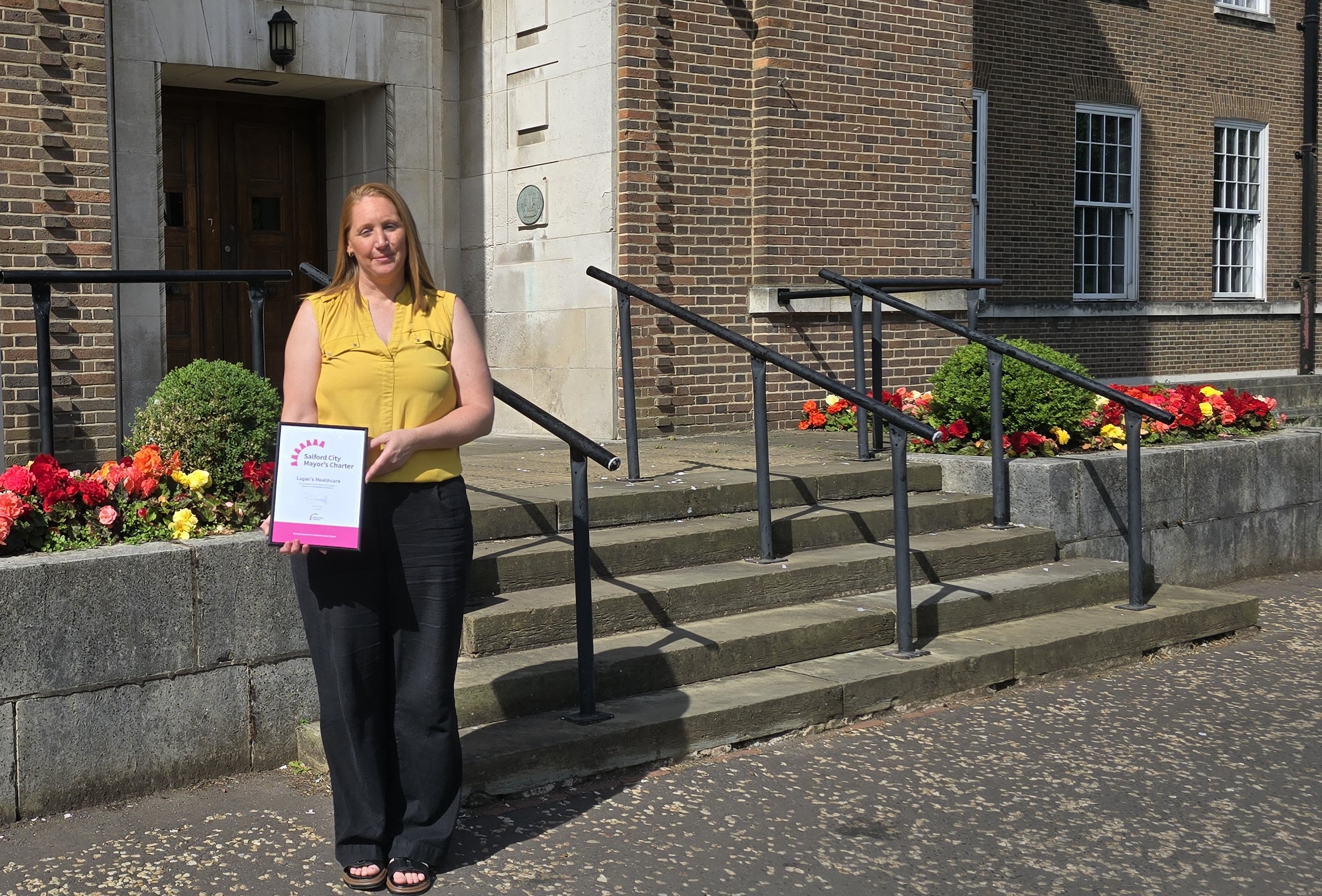 A woman stands outside smiling and holding a framed certificate. She is wearing a yellow blouse and black trousers. She stands in front of some stone steps with black railings, flowers and shrubbery.