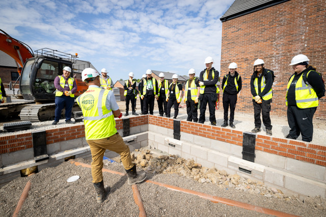 A group of young people wearing hi-vis jackets and hard hat gather around a man on a construction site who is giving them some information. The man is tall and wearing a white hard-hat, a white shirt under a hi-vis jacket that says 'Protect' on the back, brown chinos and construction boots. A digger and a house can be seen in the background.