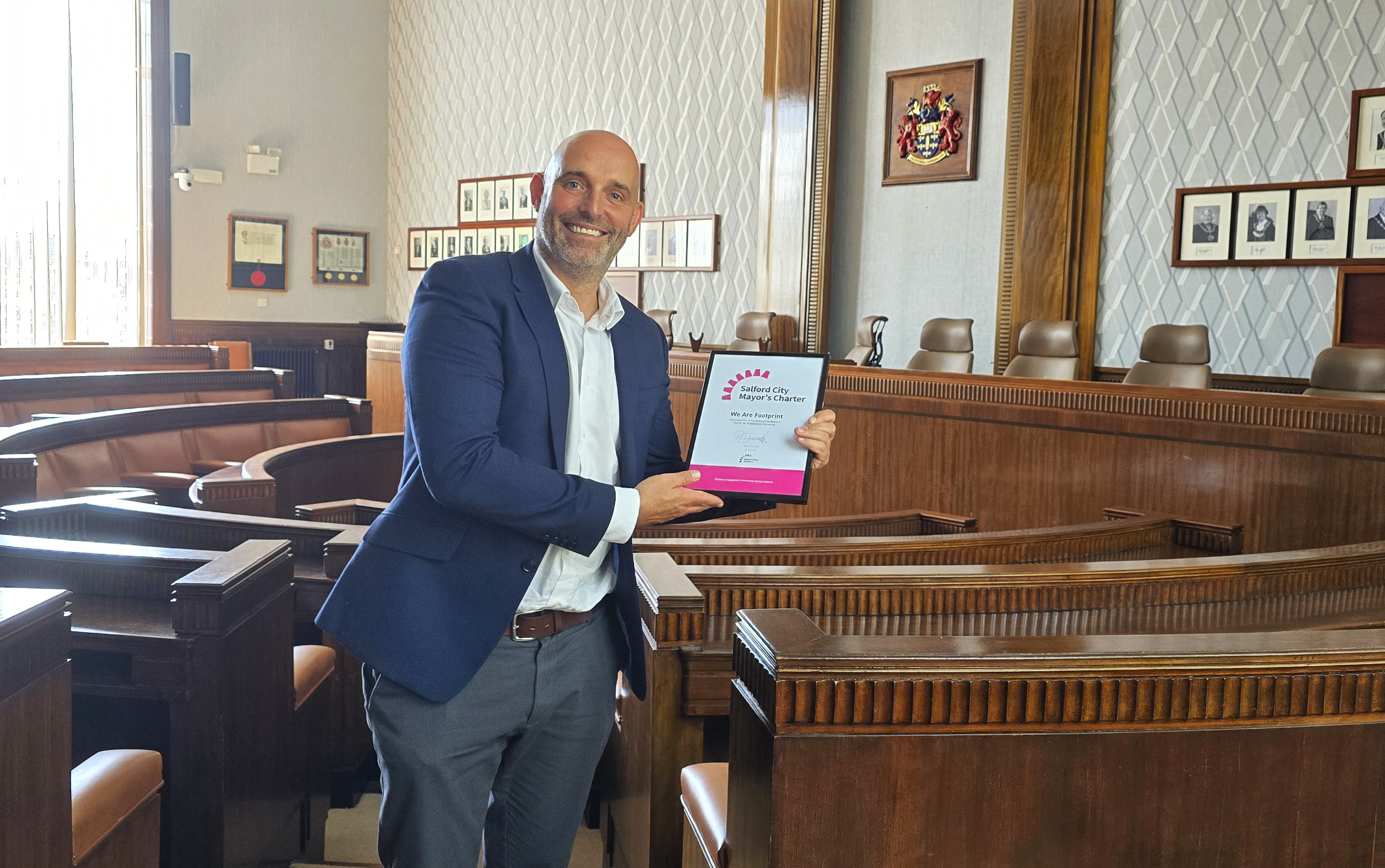 A man standing in council chamber holding an award plaque and smiling. He is wearing a navy blue blazer over a white dress shirt tucked into grey trousers. The background features wooden benches, framed pictures, and various plaques and certificates on the walls.